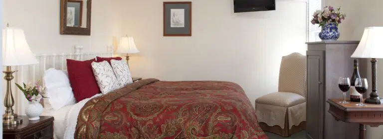 Bedroom with white walls, white painted wrought iron headboard, red and gold paisley bedding, and dark wooden furniture