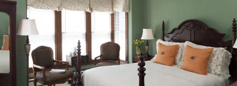 Bedroom with green walls, dark wood trim, dark wooden headboard, light colored bedding, and sitting area with antique chairs