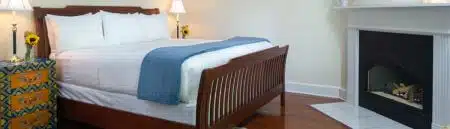 A bedroom with a bed with white and blue bedding, wood headboard, footboard and sideboards, nightstand and lamps on either side, wood floors, a blue and beige tapestry rug, plush blue armchairs with beige throw pillows and an pen book on one, an accent table in between them with glasses of white wine, and a fireplace in the corner.