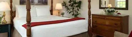 A bedroom with a canopy bed, a tan loveseat at the foot of the bed, nightstands on either side of the bed with lamps and vases of sunflowers, wood floors, cream colored walls with white trim, a tapestry rug in front of the loveseat, and a window with views of the outside grounds and green foliage.