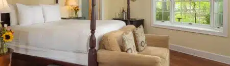 A bedroom with a canopy bed, a tan loveseat at the foot of the bed, nightstands on either side of the bed with lamps and vases of sunflowers, wood floors, cream colored walls with white trim, a tapestry rug in front of the loveseat, and a window with views of the outside grounds and green foliage.