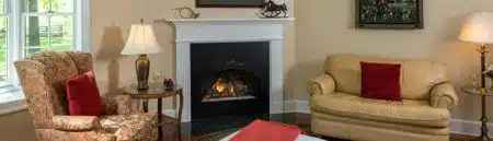 A bedroom with a bed with white and red bedding, a plush tan loveseat with red throw pillow, a stuffed tapestry armchair with red throw pillow, side table and floor lamp, and a fireplace in the corner.