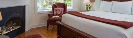 A bedroom with a bed with white and red bedding, a stuffed armchair in front of a window with views of the lush green bushes and trees outside, and a fireplace in the corner.