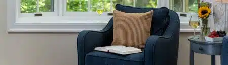 A room with a plush blue armchair with tan cushion and open book, and a side table with a glass of white wine, fresh berries and a vase with a yellow sunflower, set in front of a bay window with a glass of white wine and lush garden views.