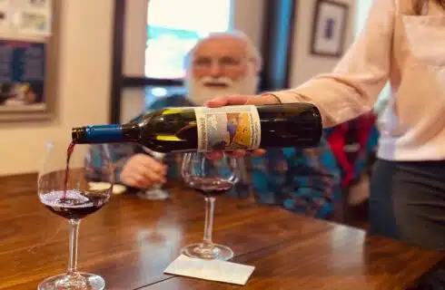 a woman pouring red wine into a wine glass with a man looking on at the table.