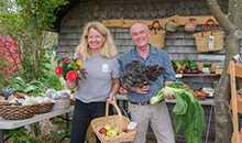 woman holding flowers and basket of apples beside man holding produce. Both are smiling. Outside Farm Store