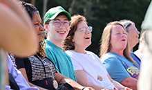 Five adults sit on a wagon bench smiling while riding through sunny summer forest